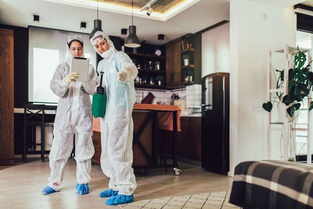Two people in hazmat suits in a kitchen, one holding a tablet, the other a sprayer.